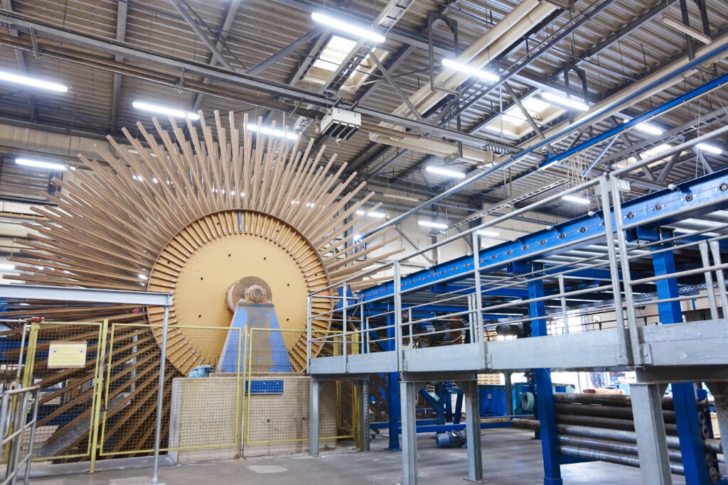 a bright blue conveyor system runs parallel to an elevated steel platform where a massive rotary storage or feeder drum used for organizing and processing long wood slats or veneers. The radial layout of the slats around the central hub creates a visually dramatic "sunburst" or "fan" pattern.